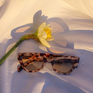 May include: Cat-eye tortoiseshell sunglasses with brown gradient lenses. A yellow daffodil with a green stem and leaves lies next to the sunglasses on a white surface.