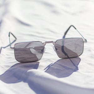 May include: Silver-framed sunglasses with gray lenses. The rectangular lenses are set in a silver frame with a bridge and temples. The sunglasses are resting on a white surface, casting a shadow.