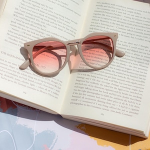 Może przedstawiać: Okulary przeciwsłoneczne z beżową oprawką i różowymi soczewkami z gradientem spoczywają na otwartej książce. Strony książki wyświetlają drukowany tekst, a okulary rzucają cień. Scena sugeruje wypoczynek i czytanie.