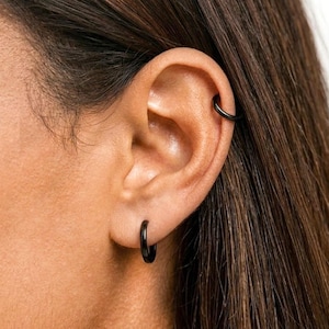 May include: Close-up of an ear adorned with black hoop earrings and a matching ear cuff. The jewelry contrasts against the skin tone and dark hair, creating a minimalist and modern aesthetic. The earrings are small and sleek.