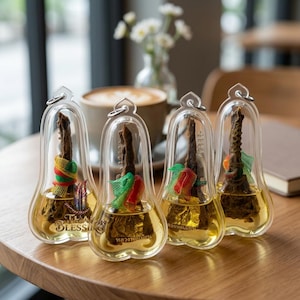 May include: Four clear, teardrop-shaped pendants containing a brown, textured object suspended in a yellow liquid. Each pendant has a colourful ribbon and the text "THA BLESSING". The pendants are on a wooden table.