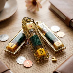 May include: Three encased dice charms with gold-coloured frames. The dice are yellow and white, with visible dots and a central red circle. The charms are displayed with coins and a notebook on a wooden surface. The text "Thai Blessings" is visible.
