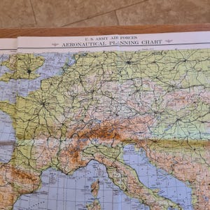 May include: A U.S. Army Air Forces Aeronautical Planning Chart featuring a detailed map of Europe. The paper map displays geographical terrain in shades of tan and green, with blue water bodies, black transportation lines, and text.