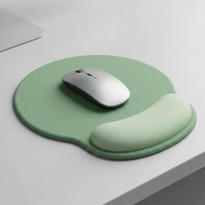 May include: A light green mouse pad with a built-in wrist rest. A silver and white wireless mouse sits on the pad. The pad is round with a curved rest for the wrist. The setup is on a white desk.