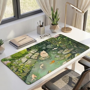 May include: A rectangular desk mat with an illustration of a pond scene, including a cat, koi carp, and lily pads. The mat has a green and brown colour scheme, with a stone path and vegetation. The desk mat is on a white desk with a gold lamp and a black chair.