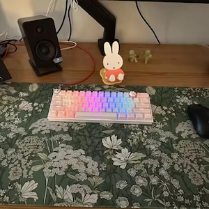 以下が含まれることがあります： レインボーカラーのバックライト付きキーを備えた白いメカニカルキーボードが、緑色の花柄のデスクマットの上に置かれています。黒いコンピューターマウスが右側にあります。2つの黒いスピーカーとラップトップも表示されています。