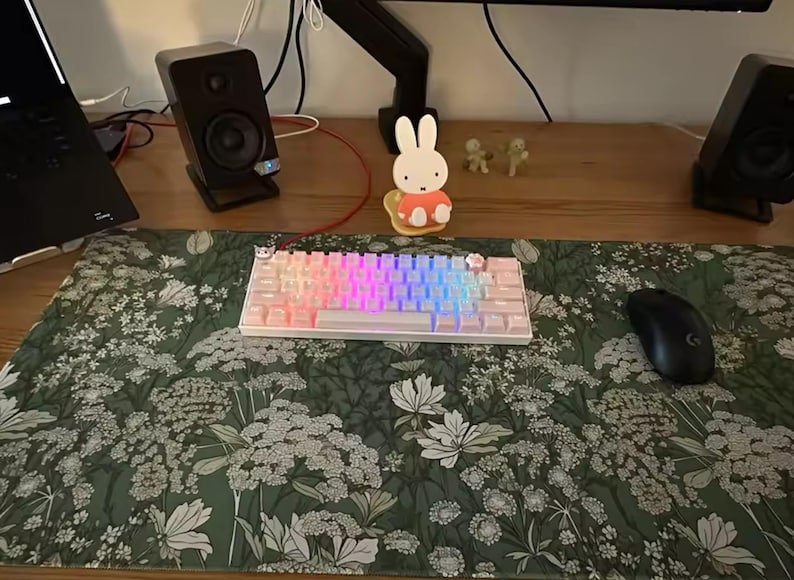 以下が含まれることがあります： レインボーカラーのバックライト付きキーを備えた白いメカニカルキーボードが、緑色の花柄のデスクマットの上に置かれています。黒いコンピューターマウスが右側にあります。2つの黒いスピーカーとラップトップも表示されています。