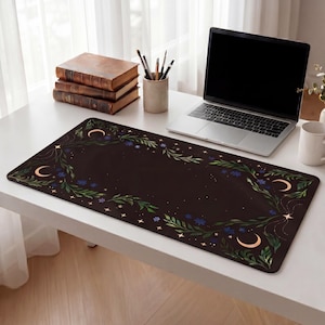 May include: A dark brown desk mat with a celestial design featuring crescent moons, stars, and floral elements. The mat is rectangular and sits on a white desk, alongside a laptop, books, and a mug.