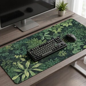 Op de afbeelding: Een rechthoekige bureaumat met een weelderig groen botanisch ontwerp, met verschillende bladvormen en -tinten. Een zwart toetsenbord en muis liggen op de mat, die een houten bureau bedekt. Een computermonitor en een kleine potplant zijn ook zichtbaar.