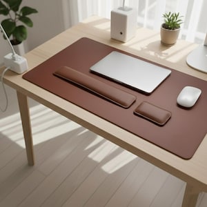 Pode incluir: Um tapete de mesa castanho com apoio de pulso e tapete de rato a condizer. Um laptop prateado e um rato branco estão no tapete. A mesa é feita de madeira clara. Uma régua de tomadas branca e uma pequena planta em vaso também são visíveis.