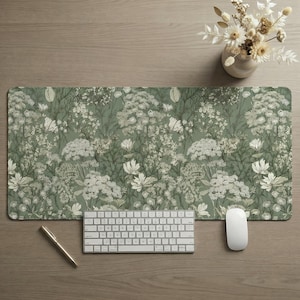 May include: A rectangular desk mat with a floral pattern in shades of green and white. A white keyboard, mouse, and a gold pen are on the mat. A small white vase with dried flowers sits in the upper right corner.