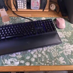 以下が含まれることがあります： 黒いRazerメカニカルキーボードとリストレストが、花柄のデスクマットの上にあります。ピンクのワイヤレスマウスがキーボードの右側にあります。デスクマットは、白い花が咲く緑色の背景です。