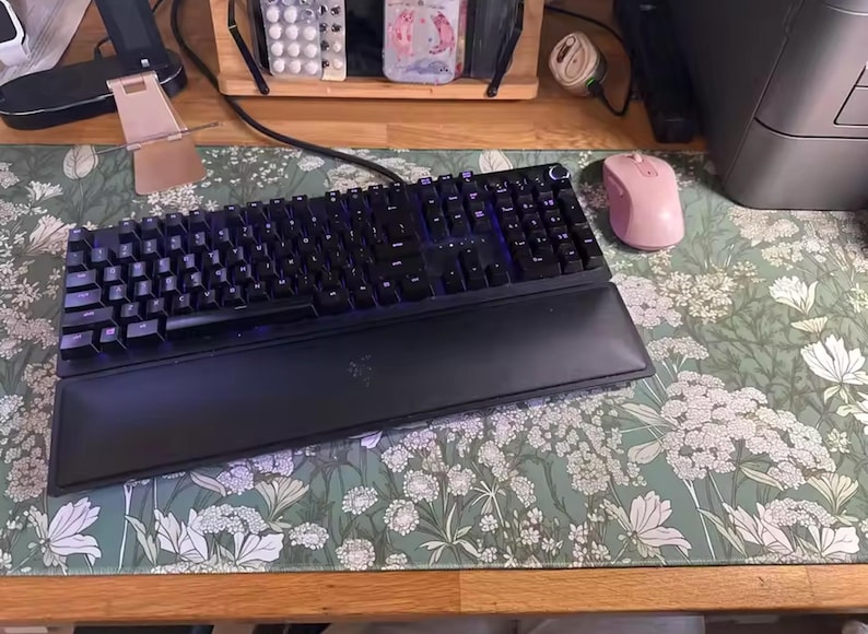 以下が含まれることがあります： 黒いRazerメカニカルキーボードとリストレストが、花柄のデスクマットの上にあります。ピンクのワイヤレスマウスがキーボードの右側にあります。デスクマットは、白い花が咲く緑色の背景です。