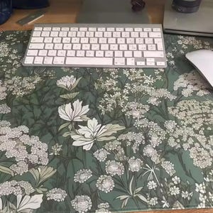 以下が含まれることがあります： 緑色の背景に白と薄緑色の花と葉が描かれた、フローラル柄のデスクマット。白いキーボードとマウスが上にあります。デスクの表面を保護するように設計されています。