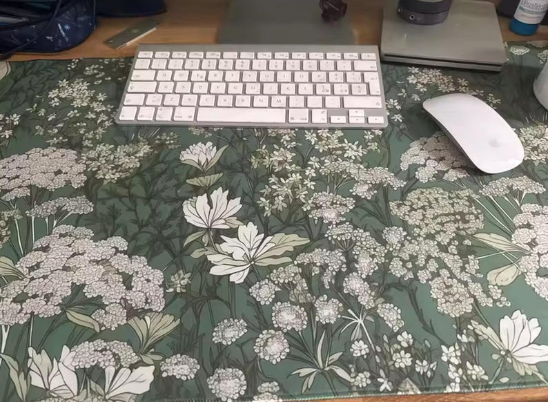 以下が含まれることがあります： 緑色の背景に白と薄緑色の花と葉が描かれた、フローラル柄のデスクマット。白いキーボードとマウスが上にあります。デスクの表面を保護するように設計されています。
