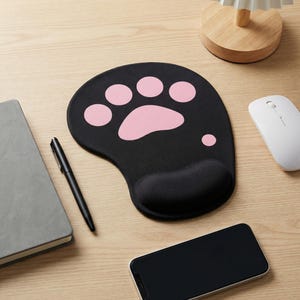 Puede incluir: Alfombrilla de ratón ergonómica negra con un diseño de huella de pata rosa. La alfombrilla tiene un reposamuñecas incorporado para mayor comodidad. Un ratón inalámbrico blanco, un bolígrafo negro, un cuaderno gris y un smartphone también están sobre el escritorio de madera.