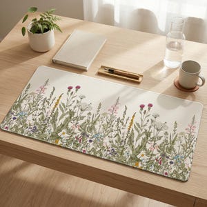 Puede incluir: Una alfombrilla de escritorio rectangular con un diseño floral de flores silvestres en tonos rosa, amarillo y azul sobre un fondo crema. La alfombrilla está sobre un escritorio de madera clara, con un cuaderno, un bolígrafo y una taza.