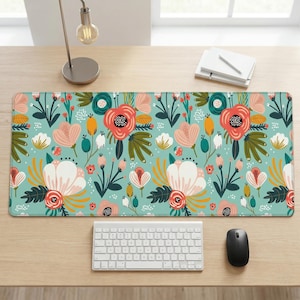 Puede incluir: Alfombrilla de escritorio rectangular con estampado floral. La alfombrilla presenta un fondo verde azulado claro con un patrón repetido de flores grandes en tonos rosa, coral y blanco, con hojas y acentos verdes. Un teclado blanco y un ratón negro están sobre el escritorio.