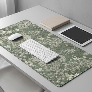 以下が含まれることがあります： 白のキーボード、マウス、タブレット、ベージュの本が付いた、緑色の花柄のデスクマット。マットは、緑色の背景に白い花と葉の繰り返し模様が特徴です。キーボードは白で長方形です。