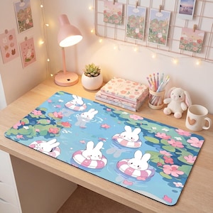 May include: A rectangular desk mat featuring a light blue water surface with pink and green lily pads and several white cartoon rabbits floating in pink and white striped inner tubes. The mat has a smooth surface and a black border edge.