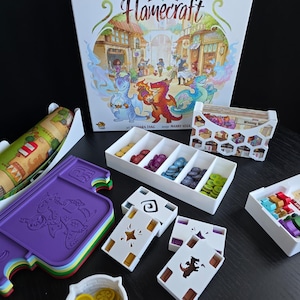 Może przedstawiać: Prezentacja gry planszowej "Flamecraft" z pudełkiem z ilustracjami smoków i sceną miejską. Widoczne są również elementy gry, takie jak karty, żetony i tace w kolorach takich jak fioletowy, zielony i żółty.