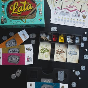 Könnte beinhalten: Ein Brettspiel mit einer türkisfarbenen Schachtel mit dem Wort "Lata" in Gelb. Das Spiel enthält Karten, Spielsteine und Holzspielsteine. Die Box-Art zeigt ein Boot und Figuren. Die Spielkomponenten sind auf einer schwarzen Oberfläche angeordnet.