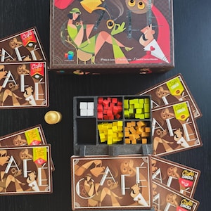 Può includere: Un gioco da tavolo con una scatola e carte marroni con la parola "Café" in un font stilizzato. L'artwork della scatola raffigura figure in verde, marrone e rosso. All'interno, ci sono pezzi di gioco in legno di vari colori, tra cui bianco, rosso, giallo e arancione.
