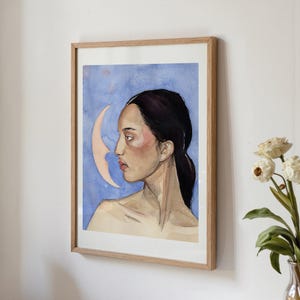 May include: Watercolor portrait of a woman in profile, framed in a light wood. The artwork features a woman with dark hair and a pale complexion against a blue background with a crescent moon. The painting has a soft, ethereal quality.