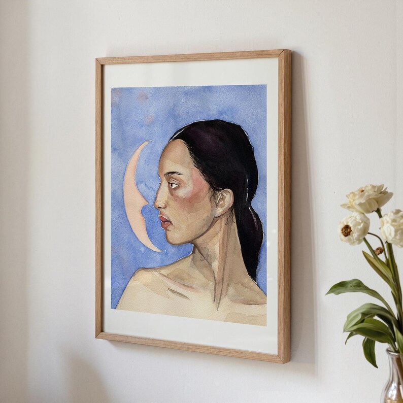 May include: Watercolor portrait of a woman in profile, framed in a light wood. The artwork features a woman with dark hair and a pale complexion against a blue background with a crescent moon. The painting has a soft, ethereal quality.