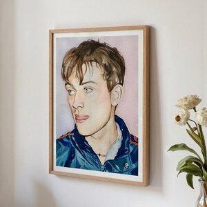 May include: Watercolor portrait of a person with brown hair and blue eyes, wearing a blue jacket with orange accents. The artwork is framed in a light-colored wooden frame and hangs on a white wall. A vase of white flowers is visible in the corner.