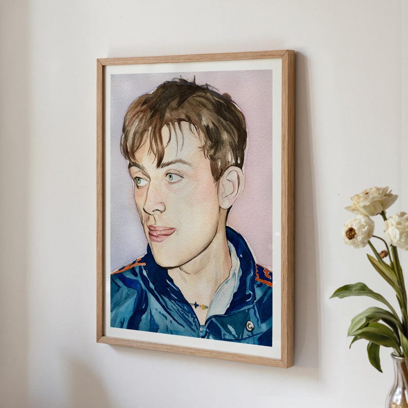 May include: Watercolor portrait of a person with brown hair and blue eyes, wearing a blue jacket with orange accents. The artwork is framed in a light-colored wooden frame and hangs on a white wall. A vase of white flowers is visible in the corner.