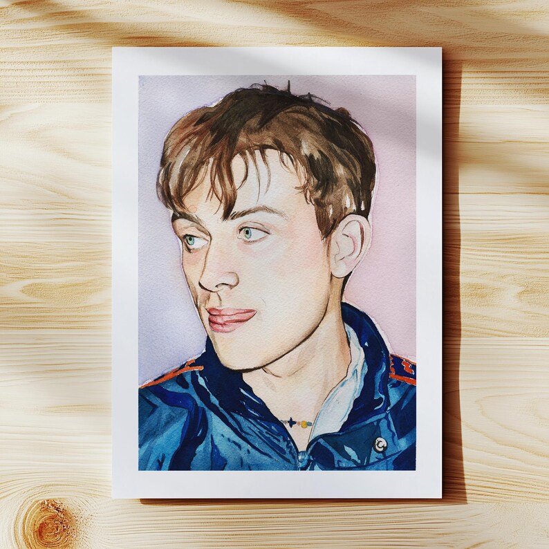 May include: Watercolor portrait of a person with brown hair and green eyes, wearing a blue jacket with orange accents. The artwork is on a white rectangular paper, set against a light purple background. A small necklace is visible.