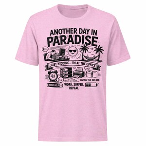 May include: Light pink t-shirt with a black and white graphic. The graphic includes the text "ANOTHER DAY IN PARADISE" and "JUST KIDDING... I'M AT THE OFFICE". Additional illustrations depict office and vacation elements.