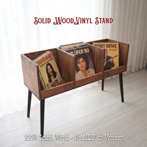 May include: A solid wood vinyl record stand with three compartments, holding vinyl records. The stand is made of dark wood and has black legs. The text "SOLID WOOD VINYL STAND" is at the top. The bottom text reads "100% Solid Wood - No MDF or Veneer".