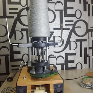 May include: A yarn winder machine made of wood and metal, with a large cone of grey yarn. The machine includes a digital display and several mechanical parts. The background shows a patterned wallpaper.