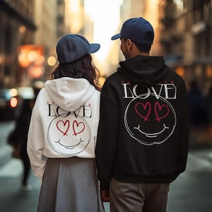 Puede incluir: Una pareja con sudaderas con capucha a juego en blanco y negro con un diseño de "LOVE". Las sudaderas tienen una carita sonriente con ojos de corazón. Ambos llevan gorras de béisbol azul marino. La imagen está tomada al aire libre.