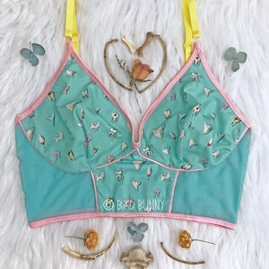 May include: A turquoise blue bra with pink trim and yellow straps. The bra is covered in a pattern of white teeth. The bra has a mesh overlay and the text "BAD BUNNY" is printed on the front.