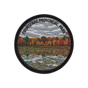 Puede incluir: Parche bordado con una vista del Parque Provincial Frontenac. El diseño incluye un lago con nenúfares, un bosque colorido y un cielo nublado. El texto "FRONTENAC PROVINCIAL PARK" está arqueado en el borde superior del parche.