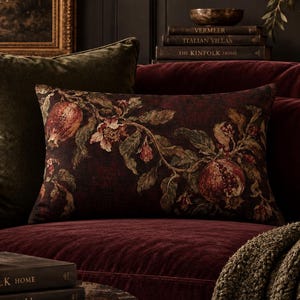 May include: A rectangular throw pillow featuring a dark burgundy background with a detailed botanical print of pomegranates, leaves, and branches. The fabric displays a rich, textured appearance with deep red and earthy green color tones.