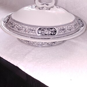 May include: A white ceramic serving dish with a lid, featuring a dark blue transferware pattern. The design includes intricate scrollwork, floral motifs, and classical urns. The lid has a central loop handle, and the base has a wide rim.