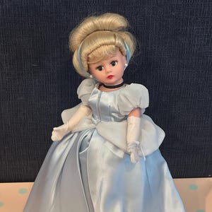 May include: A vintage doll dressed in a light blue satin gown with a matching bodice and puffed sleeves. The doll has blonde hair styled in an updo, a black choker, and white gloves. The doll's face has painted features, including rosy cheeks and red lips.