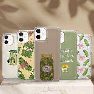 May include: Five clear smartphone cases featuring pickle-themed designs. One shows a jar held by hands, another has a yellow background with pickles and pink bows, one features a jar labelled Dill Pickles, one has green text, and one has a pickle pattern.