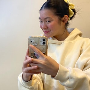 May include: A person wearing a cream-colored hoodie and holding a clear phone case with a Snoopy design. The person has a yellow crocheted hair accessory. The background is a plain, off-white wall.