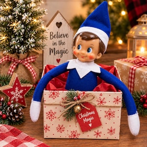 May include: A doll with a blue hat and blue outfit sits inside a square box decorated with red snowflakes. The box features a red tag with the text Christmas Magic, a small gold bell, and a twine bow. The doll has white cuffs and collar.