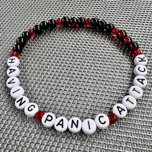 May include: A circular beaded bracelet featuring alternating black and translucent red spherical beads, interspersed with white cylindrical beads that spell out the phrase HAVING PANIC ATTACK in black capital letters around the circumference.