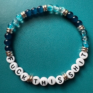 Peut inclure: Un bracelet de perles bleues et transparentes, entrecoupées d'entretoises argentées. Des perles blanches épellent "FUCK THIS SHIT" sur le pourtour. Le bracelet est présenté sur une surface turquoise.