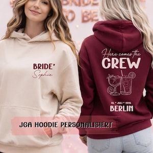 Op de afbeelding: Twee vrouwen dragen gepersonaliseerde hoodies. Eén is beige met "Bride" en een naam, de andere is bordeauxrood met "Here comes the CREW" en een stad en datum. De achtergrond heeft ballonnen en een "Bride to Be" banner.
