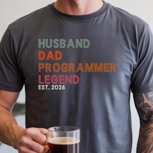 Könnte beinhalten: Dunkelgraues T-Shirt mit dem Text "HUSBAND DAD PROGRAMMER LEGEND EST. 2026" in Türkis, Orange, Braun und Rot. Die Person hält einen transparenten Glasbecher mit einem dunklen Getränk.