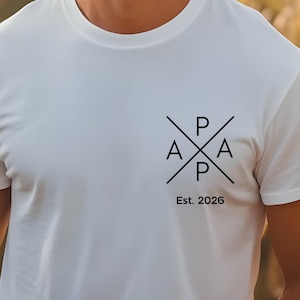 Könnte beinhalten: Weißes T-Shirt mit einem schwarzen Grafikdesign auf der Brust. Das Design zeigt die Buchstaben "P", "A" und "Est. 2026" in einem Kreuzmuster angeordnet. Das Shirt besteht aus einem weichen Material.