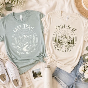 Könnte beinhalten: Zwei T-Shirts mit Outdoor-Grafiken. Eines ist salbeigrün mit dem Text "LAST TRAIL before the veil". Das andere ist cremefarben mit dem Text "BRIDE-TO-BE WILD & FREE". Weiße Turnschuhe, eine Wasserflasche und ein Hut sind ebenfalls zu sehen.